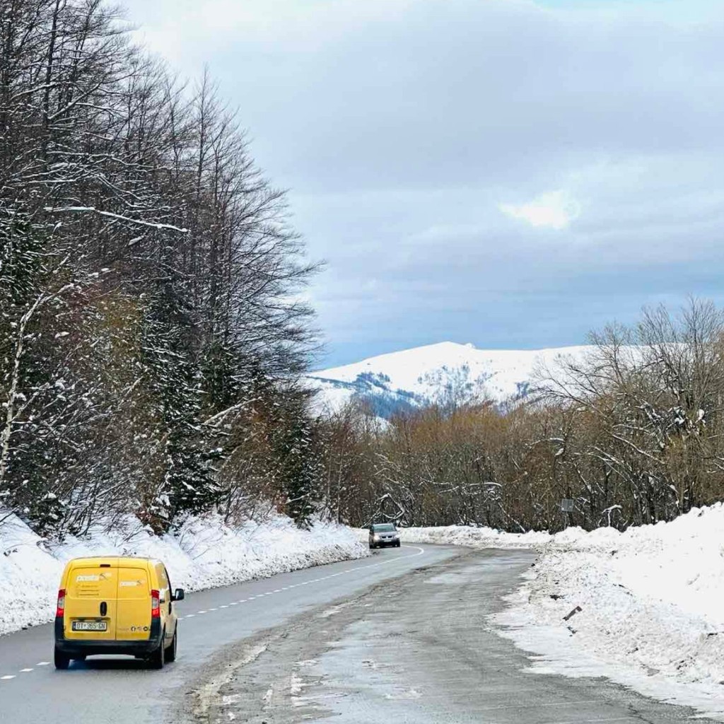 Furgon i verdhë i Postës me logo në rrugë dimri në peizazh malor.